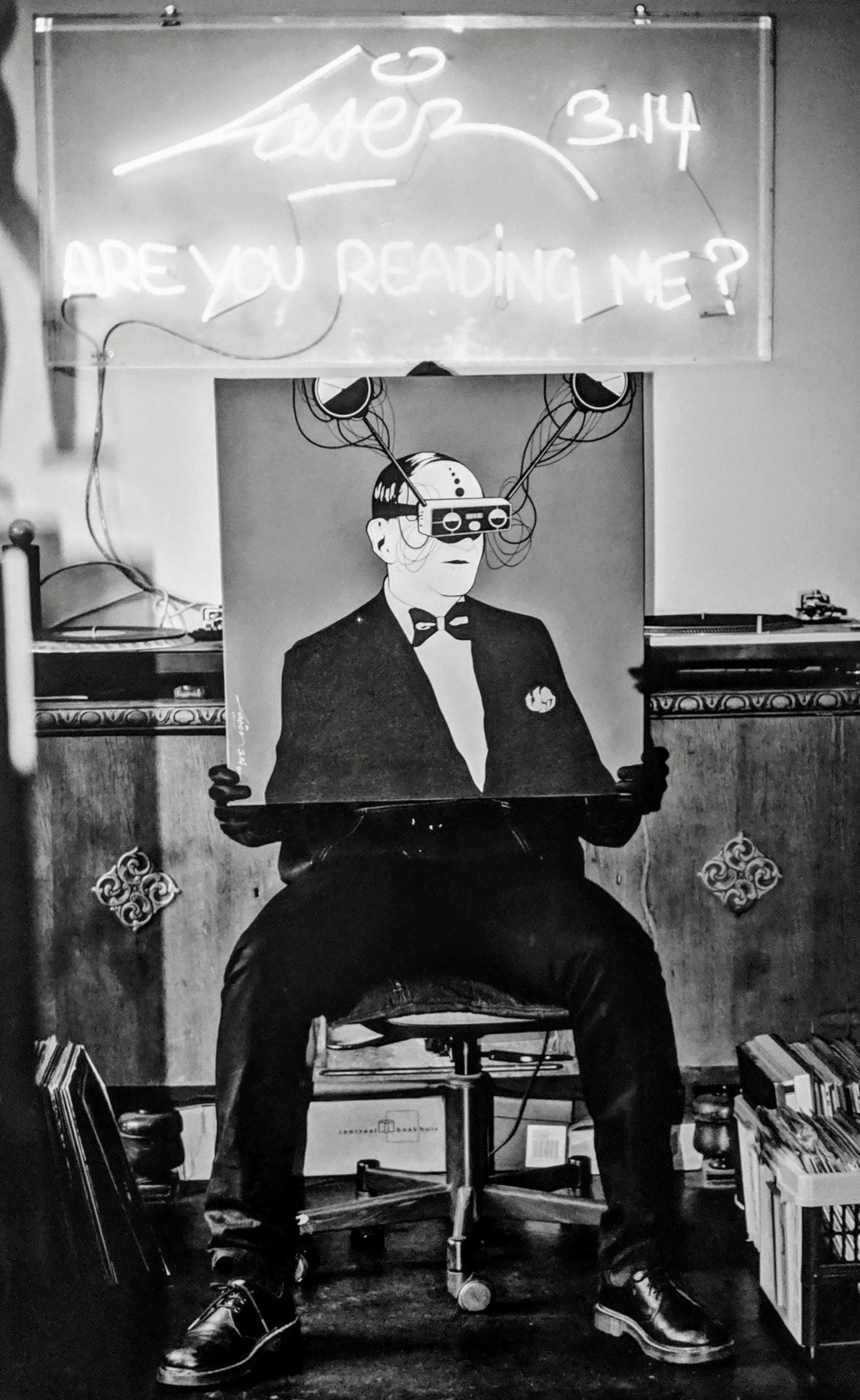 Artistic photo of a man man holding a painting with a neon light above him reading "Laser 3.14 - Are You Reading Me?"